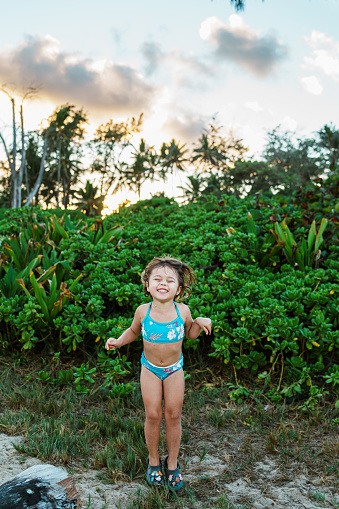 A happy and excited four year old girl of Eurasian descent stands outside in front of lush greenery, jumping up and down.