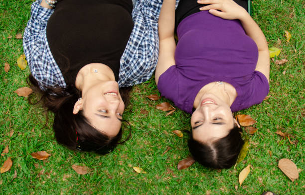 A flat lay shot of two young women with light skin having a heartfelt conversation while lying on the green grass in a sunny public park. The image captures a warm, friendly moment in a natural outdoor setting, perfect for themes of friendship, relaxation, mindfulness, and outdoor leisure.
