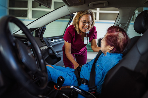 A kind occupational therapist of Hawaiian and Finnish descent smiles as she helps a female geriatric patient get seated and buckled into the passenger seat of a car.