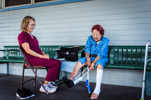 A geriatric woman of Asian descent who has limited mobility sits outside with her occupational therapist and smiles as she practices using a device designed to help her get her shoes and sock on independently.