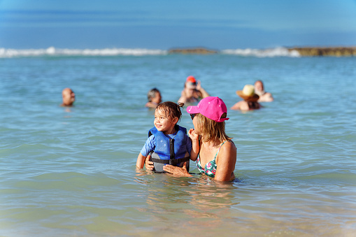 An active Eurasian senior woman playfully interacts with her three year old granddaughter as they swim together in the ocean. The cheerful little girl is wearing a lifejacket.