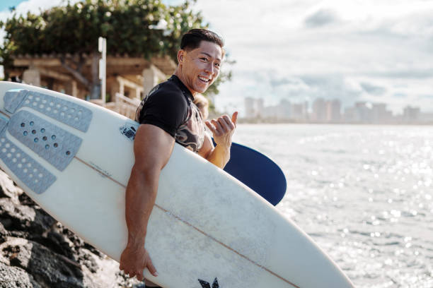 Portrait of a young and fit male surfer of Hawaiian and Japanese descent smiling and holding the shaka sign while walking on a rocky beach with his surfboard with Honolulu, Hawaii visible in the background.