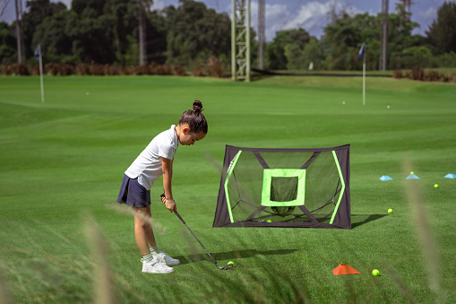 Kindergarten aged girl of Asian ethnicity stands in an athletic stance, practicing her golf swing into a set up net in front of her.