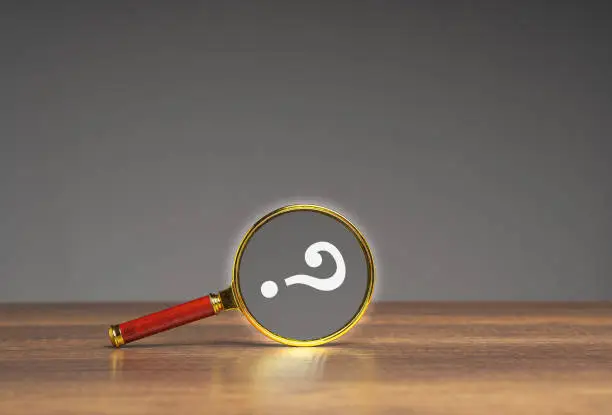 A magnifying glass with a question mark on a wooden table against a gray background. Space for text.