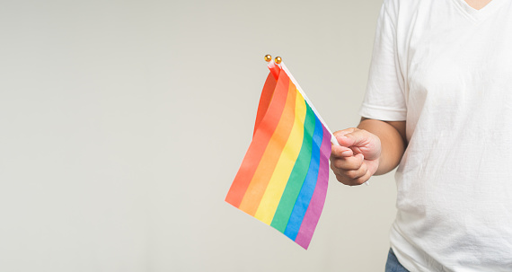 Close-up of a hand holding a rainbow flag while standing on a gray background. Calling on all people to support and respect diversity, gender, and human rights and celebrate LGBTQ Pride month.