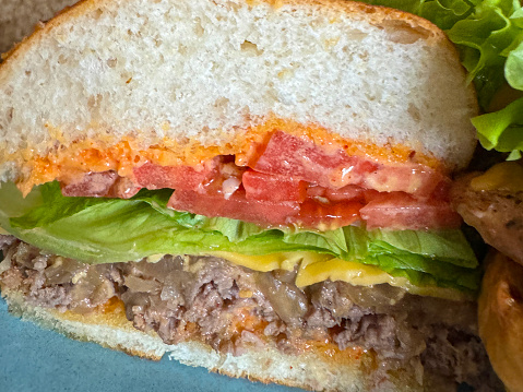 Stock photo showing close-up view of blue plate containing half a cheeseburger in a brioche bun, minced beef patty topped with melting cheese slice, lettuce and sliced tomato.