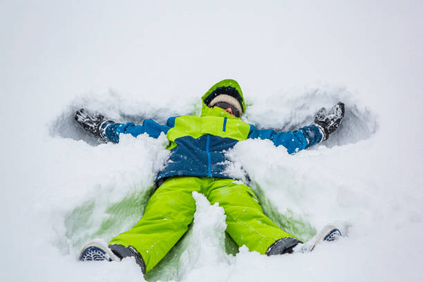 Person wearing a green beanie and winter clothing is lying face down, exhausted in deep snow, needing a break during a cold outdoor activity on a winter day in the forest