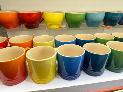 Stock photo of close-up view of retail display shelves containing rows of pottery mugs and cups in rainbow colours in shades of red, orange, yellow, blue and green glazes, glazed and fired in kiln.