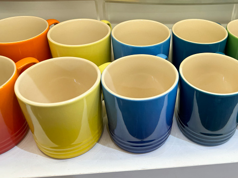 Stock photo of close-up, elevated view of retail display shelves containing rows of pottery mugs and cups in rainbow colours in shades of red, orange, yellow, blue and green glazes, glazed and fired in kiln.