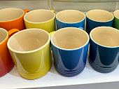 Close-up image of rows of pottery rainbow colour mugs, display shelves of group coffee / tea cups in gradient shades of red, orange, yellow, blue and green glazes, focus on foreground, elevated view