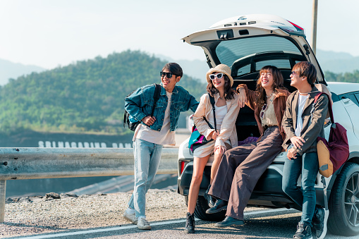 Group of Asian generation z people friends sitting on car trunk looking beautiful nature of countryside. Man and woman friendship enjoy and fun outdoor lifestyle road trip on summer holiday vacation.