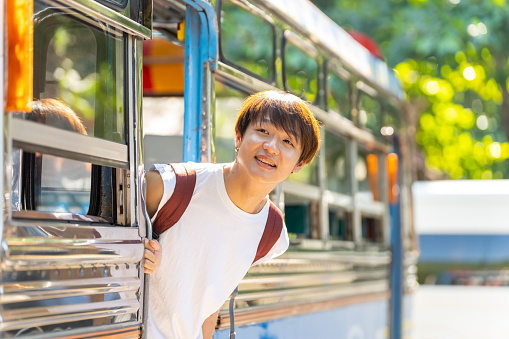 Young Asian man enjoy and fun outdoor lifestyle road trip travel nature by public transportation on summer holiday vacation. Happy guy standing on the bus looking beautiful nature of countryside.