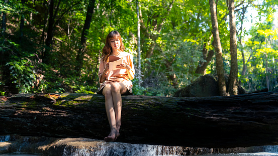 Happy young Asian woman enjoy and fun outdoor lifestyle travel nature forest mountain on summer holiday vacation. Attractive girl sitting on tree trunk and writing journey diary at nature waterfall.