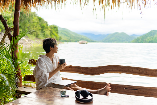 Asian Non-binary person relaxing after remote working corporate business on laptop computer on lake house balcony. People enjoy outdoor lifestyle work and travel nature on summer holiday vacation.