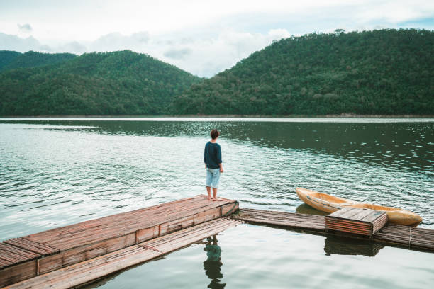 Young Asian man relax and enjoy outdoor lifestyle travel forest mountain on summer holiday vacation. Generation z guy relaxing on lake house balcony looking beautiful nature and breathing fresh air
