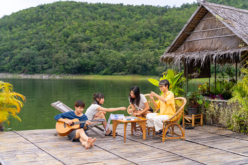 Group of Young Asian man and woman enjoy outdoor lifestyle travel nature forest mountain on summer holiday vacation. Happy generation z people friends playing guitar and doing hobbies at lake house balcony.