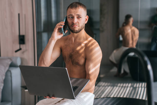 istock Multitasking man in a towel working on a laptop while talking on the phone, in a modern home environment with a mirror reflection 1899359108