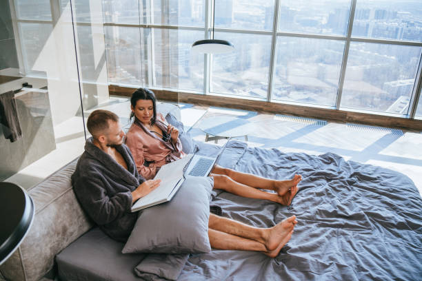 istock Man and woman in robes engaging with a book and laptop, comfortable work-from-home setting with a cityscape view 1899359123