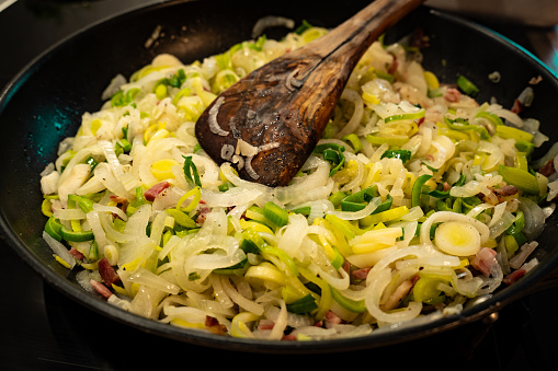 Sauté leeks, onions and bacon, ingredients for a quiche lorraine in a pan.  Close-up with wooden cooking spoon