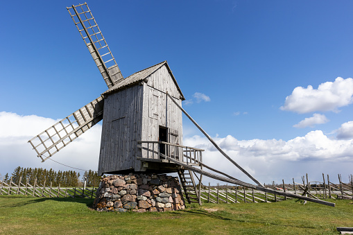 A wooden windmill on a stone foundation with a wooden fence against a blue sky background