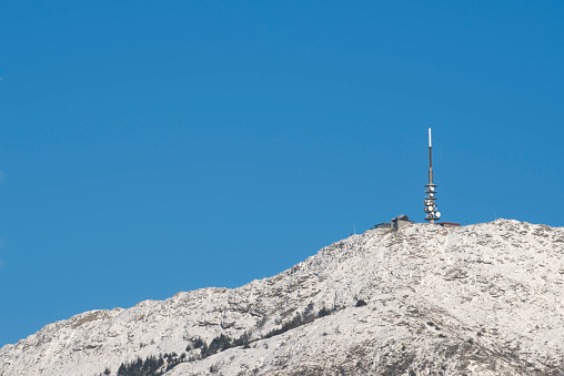 A snow covered Mount Ulriken on a sunny winter day with clear blue sky. Mount Ulriken is the highest of the seven mountains that surround the city of Bergen on the western coast of Norway. It has an altitude of 643 metres above sea level and can be reached by foot or by a cable car. At the top there is a TV tower and a restaurant.