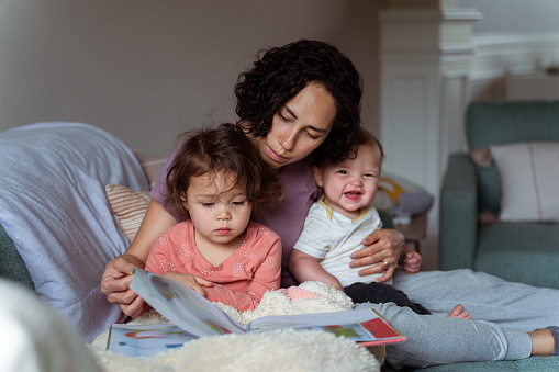 A loving multiracial mother of Asian and Pacific Islander descent holds her cheerful 8 month old son on her lap and reads a storybook to her sleepy preschool age daughter while snuggled up on the living room sofa. The family is enjoying a relaxing and cozy morning together at home.