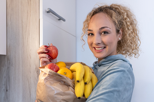 A smiling girl came home from shopping, holds a paper bag full of healthy fruits (bananas, apples) and vegetables (tomatoes) in her hand, stands in front of the refrigerator, puts the fruits in the refrigerator,
