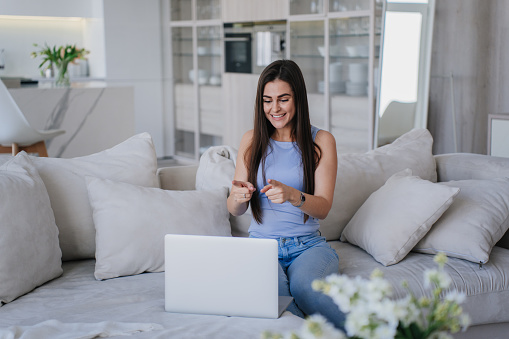 Delighted woman in blue top and jeans using a laptop on a cozy couch, surrounded by plush pillows and homey decor.