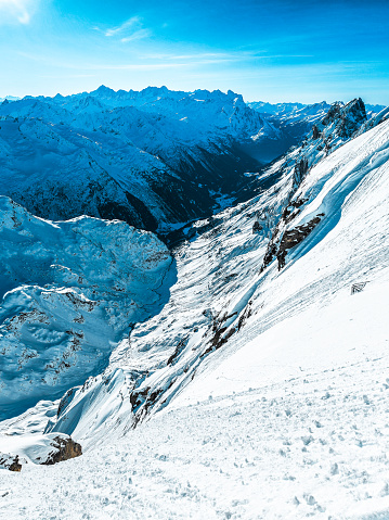 On Titlis mountain, a spectacular winter landscape presents panoramic views over the Swiss Alps. It's a very clear, sunny day, enhancing visibility so that one can see far into the distance. The scene is dotted with numerous mountain tops, each uniquely shaped and covered in glistening snow. The vast snowy landscapes stretch out, creating a serene and awe-inspiring atmosphere, with the bright sunlight casting sharp shadows and illuminating the pristine beauty of the alpine terrain.