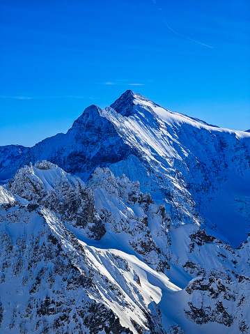 On Titlis mountain, a spectacular winter landscape presents panoramic views over the Swiss Alps. It's a very clear, sunny day, enhancing visibility so that one can see far into the distance. The scene is dotted with numerous mountain tops, each uniquely shaped and covered in glistening snow. The vast snowy landscapes stretch out, creating a serene and awe-inspiring atmosphere, with the bright sunlight casting sharp shadows and illuminating the pristine beauty of the alpine terrain.