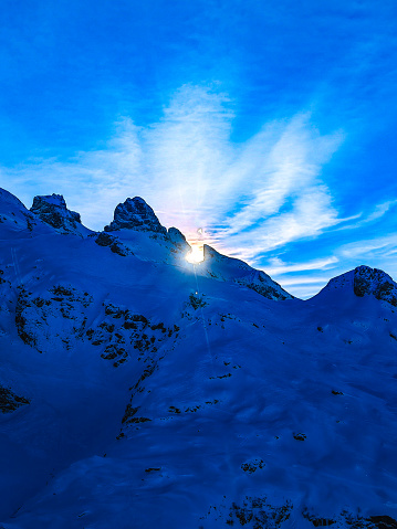 On Titlis mountain, a spectacular winter landscape presents panoramic views over the Swiss Alps. It's a very clear, sunny day, enhancing visibility so that one can see far into the distance. The scene is dotted with numerous mountain tops, each uniquely shaped and covered in glistening snow. The vast snowy landscapes stretch out, creating a serene and awe-inspiring atmosphere, with the bright sunlight casting sharp shadows and illuminating the pristine beauty of the alpine terrain.