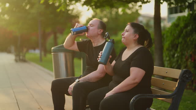 Senior couple taking a rest and drinking water while sitting at park bench in public park