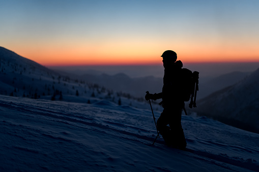 silhouette of a tourist man with hiking equipment stands on a snowy mountain slope against the background of the sky and sunset
