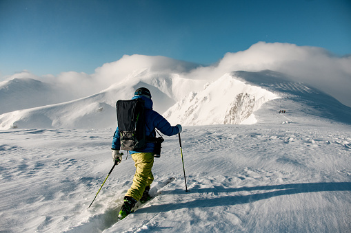 rear view of male skier with hiking equipment walking at deep snow on mountain slope. Beautiful winter landscape and blue sky at background