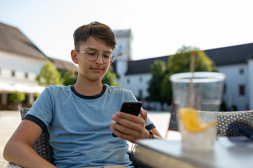 A teenager stares blankly at his phone while walking around the city, sit at the table.