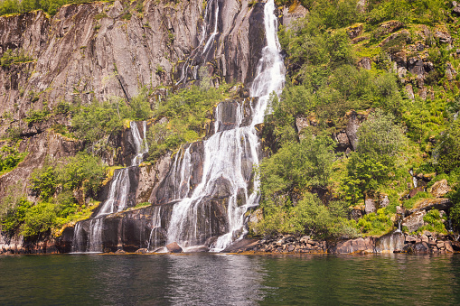 A vintage-toned photo captures a majestic waterfall cascading into Trollfjorden, Lofoten, Norway, surrounded by lush greenery and sun-kissed cliffs