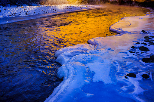 Golden Sunrise Light Cold River Landscape - Scenic views along mountain river in winter with golden light contrasting with cool blue shadows. Ice and icy reflections in water.