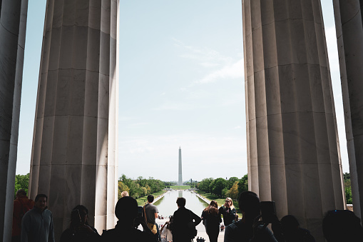 Standing inside the Lincoln Memorial facing out, the Washington Monument can be seen across the Reflecting Pool on the horizon in the distance. The image is framed on both sides by large marble columns and the silhouettes of tourists.