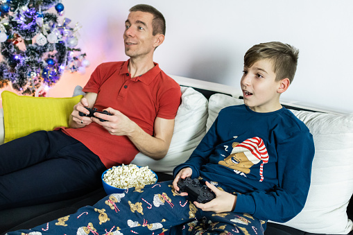 Happy father and son dressed in pajamas are playing video games next to decorated Christmas tree.