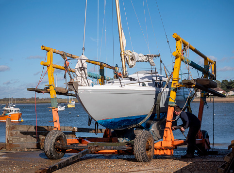 A sailing boat has been hoisted out of the River Deben estuary at Felixstowe Ferry in Suffolk, Eastern England, and a man works with a jet-washer to remove weed, grime and debris from its hull. The hamlet of Bawdsey is on the opposite bank.
