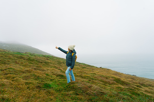Latin woman walking up a hill and pointing to a part of the landscape on the Irish coast on a foggy day