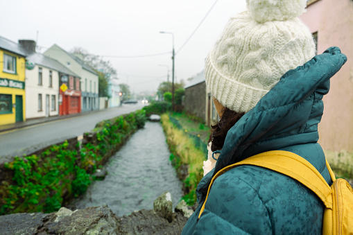 Woman seen from behind wearing knit hat and backpack with water canal passing through a small town in Ireland