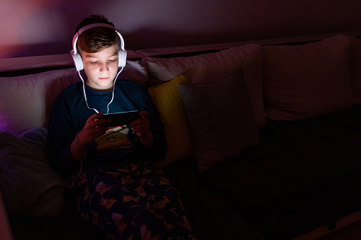 Happy boy dressed in pajamas with headphones is playing video games next to decorated Christmas tree.