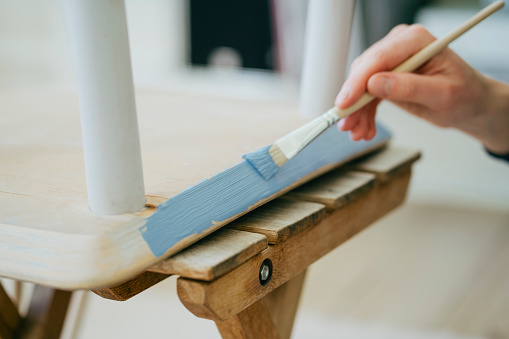 DIY transformation: Woman's hands skillfully painting an antique chair in blue with a paint brush.