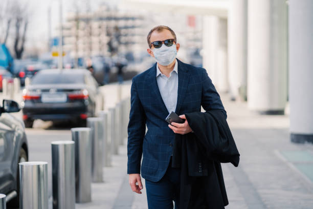 istock Professional man in a blue checkered suit walks confidently in the city, wearing a protective face mask and sunglasses, holding a phone. 1900215802