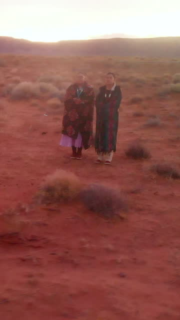 Fly Away Aerial Drone Footage Two Navajo Sisters Standing in Desert in Monument Valley Utah, Tribal Park in Traditional Dress at Dusk Under Dramatic Sunset Sky