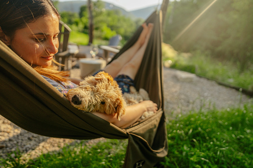 Photo of a young woman swinging in a hammock with her dog and enjoying a beautiful sunny day