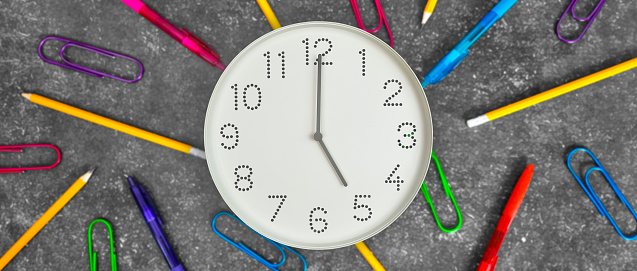 Clock  with white clock face on dark background with pencils, pens and paper clips. Concept of study, work, deadline, schedule. Time management.