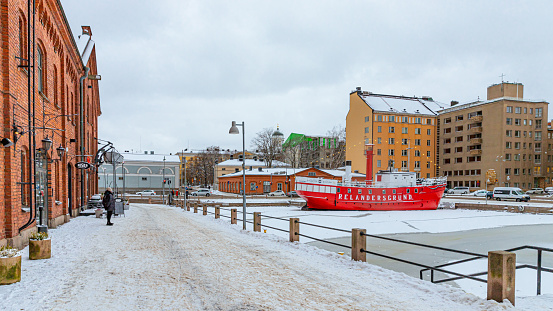Helsinki, Finland. December 28th, 2023. A red vessel converted into a restaurant stuck in the ice of the Baltic Sea and a single tourist viewing the cityscape.