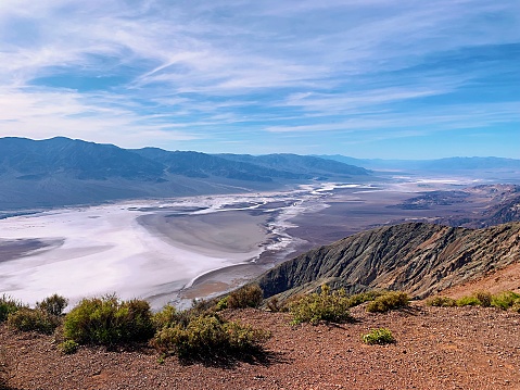 The Valley in Death Valley, California with bright red sand and small desert bushes growing on the side. Below are the large salt flats. There are mountains in the back that are very tall. There are patterns on the rock. The sky is blue with clouds.